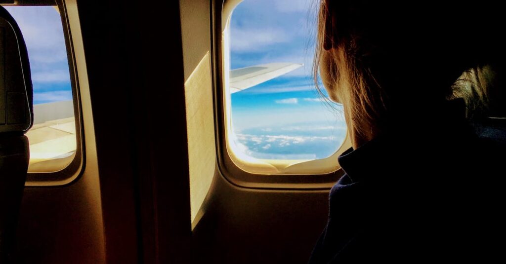 Silhouette of a woman looking out an airplane window at clouds and blue sky.