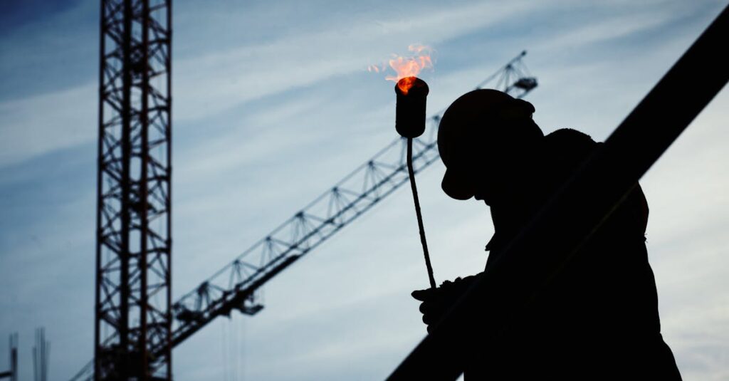 Silhouette of a construction worker using a blowtorch at a building site against a crane-filled skyline.