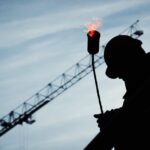 Protecting the Pulse of Our Workforce Silhouette of a construction worker using a blowtorch at a building site against a crane-filled skyline.