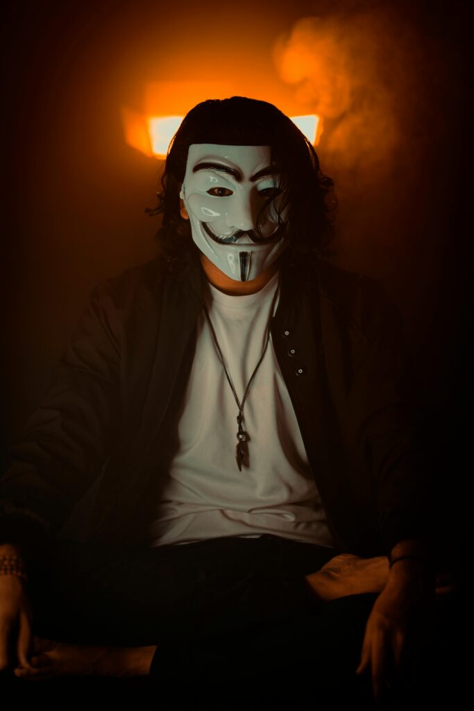 A person wearing a Guy Fawkes mask sits in a dark, smoky room with warm lighting.