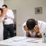 Despairing worker at office desk as colleagues gossip in background illustrating workplace bullying.