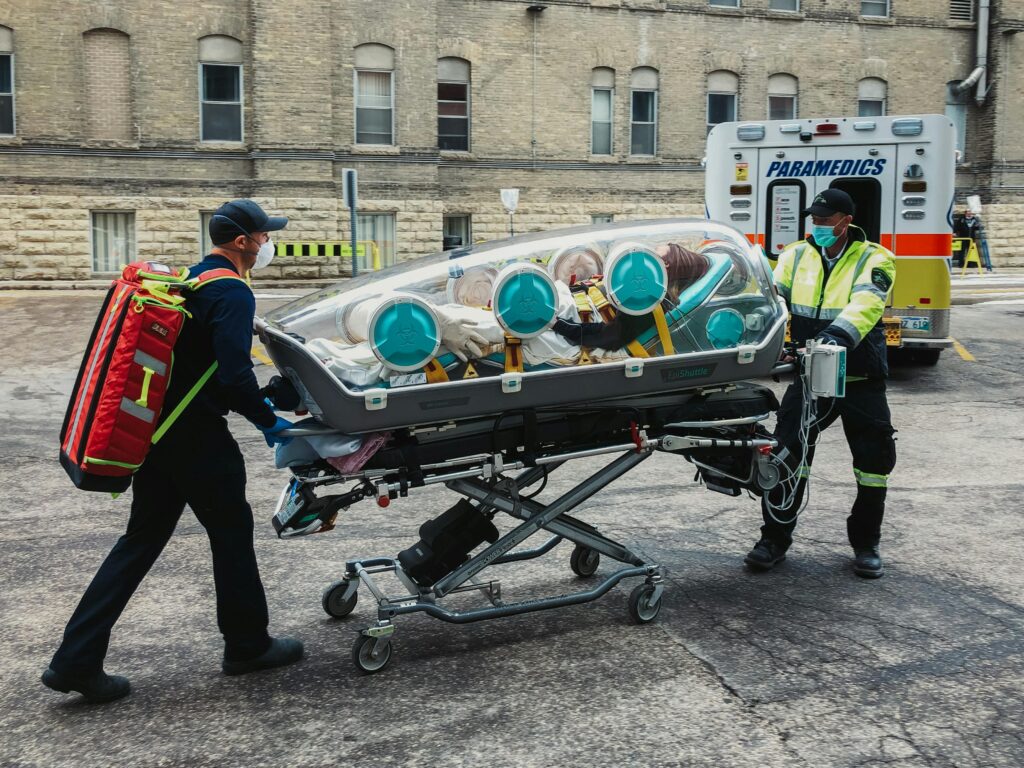 Emergency paramedics transport a patient in an isolation pod outside a medical facility.