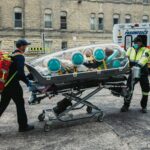 Emergency paramedics transport a patient in an isolation pod outside a medical facility.