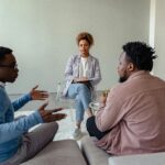 A therapist mediates a session with two men on a couch discussing mental health.