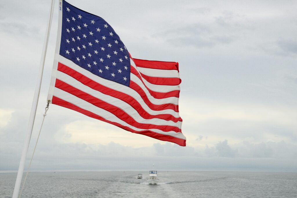 A vibrant American flag flutters on a boat with a vast ocean and cloudy sky backdrop.
