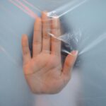 A close-up of a hand pushing against a translucent plastic sheet, suggesting themes of confinement or barrier.